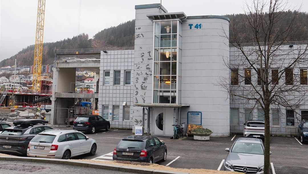 En toetasjes grå kontorbygning merket "T 41" med parkerte biler foran og synlige byggearbeider til venstre.
