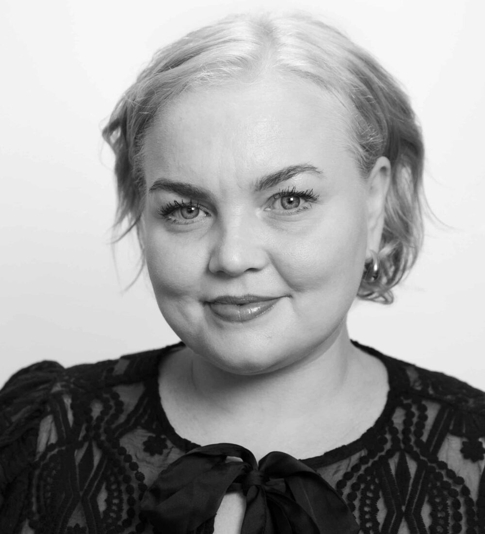 Michelle Sjo Lønne En kvinne med lyst hår og blondetopp smiler lett mens hun ser inn i kameraet mot en ensfarget bakgrunn.