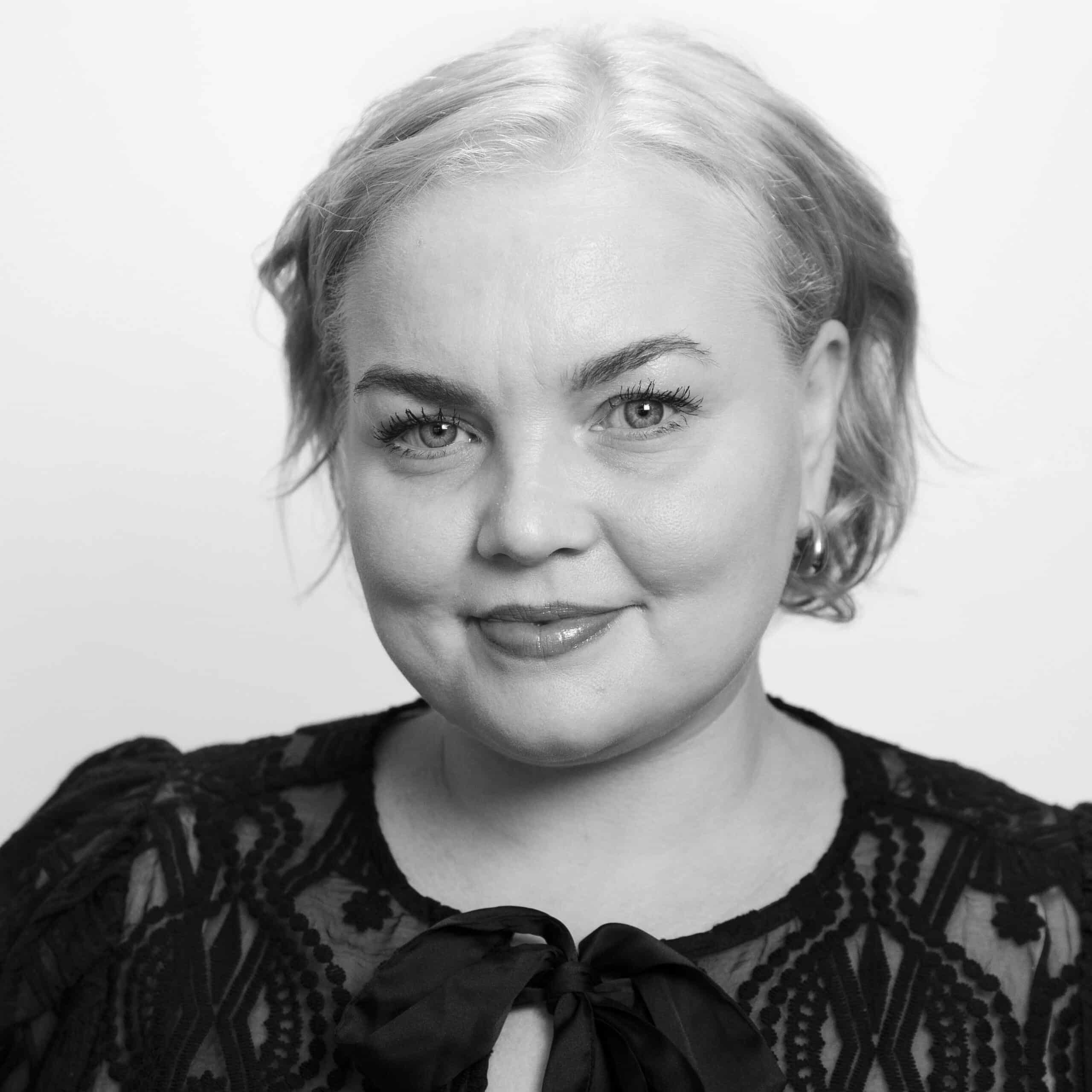 En kvinne med lyst hår og blondetopp smiler lett mens hun ser inn i kameraet mot en ensfarget bakgrunn.