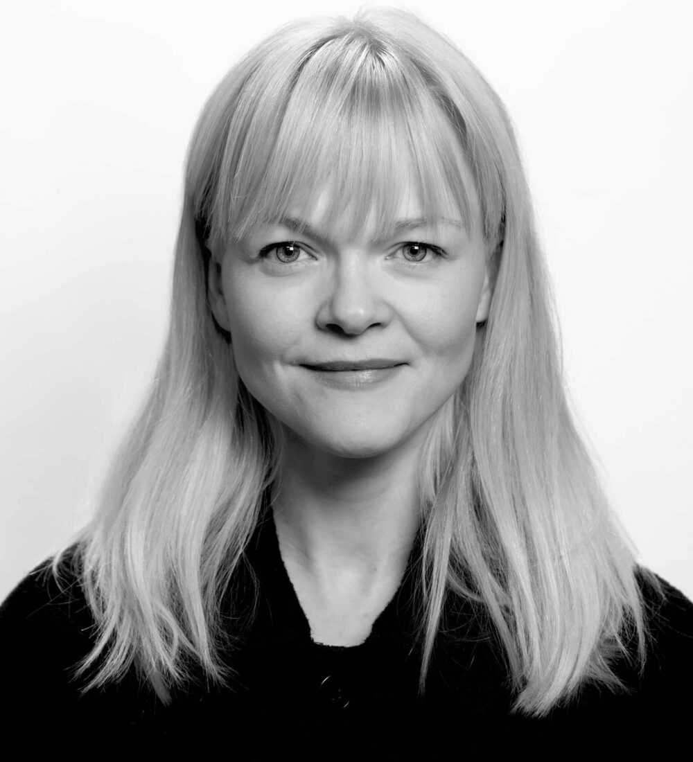 En kvinne med rett, lysblondt hår og lugg, iført en mørk topp, står vendt mot kameraet og smiler lett mot en enkel, hvit bakgrunn.