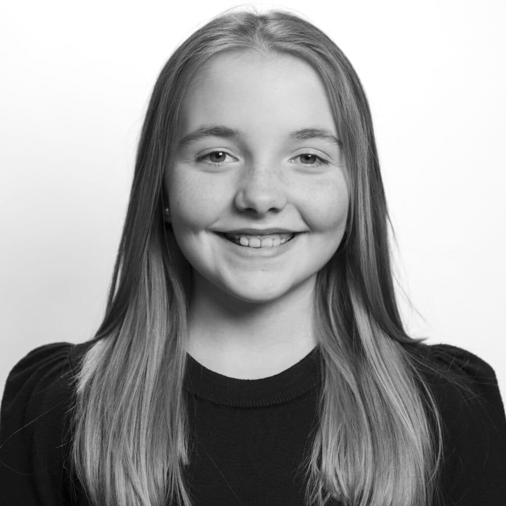 Svart-hvitt portrett av en ung jente med langt, glatt hår som smiler til kameraet mot en ensfarget bakgrunn.