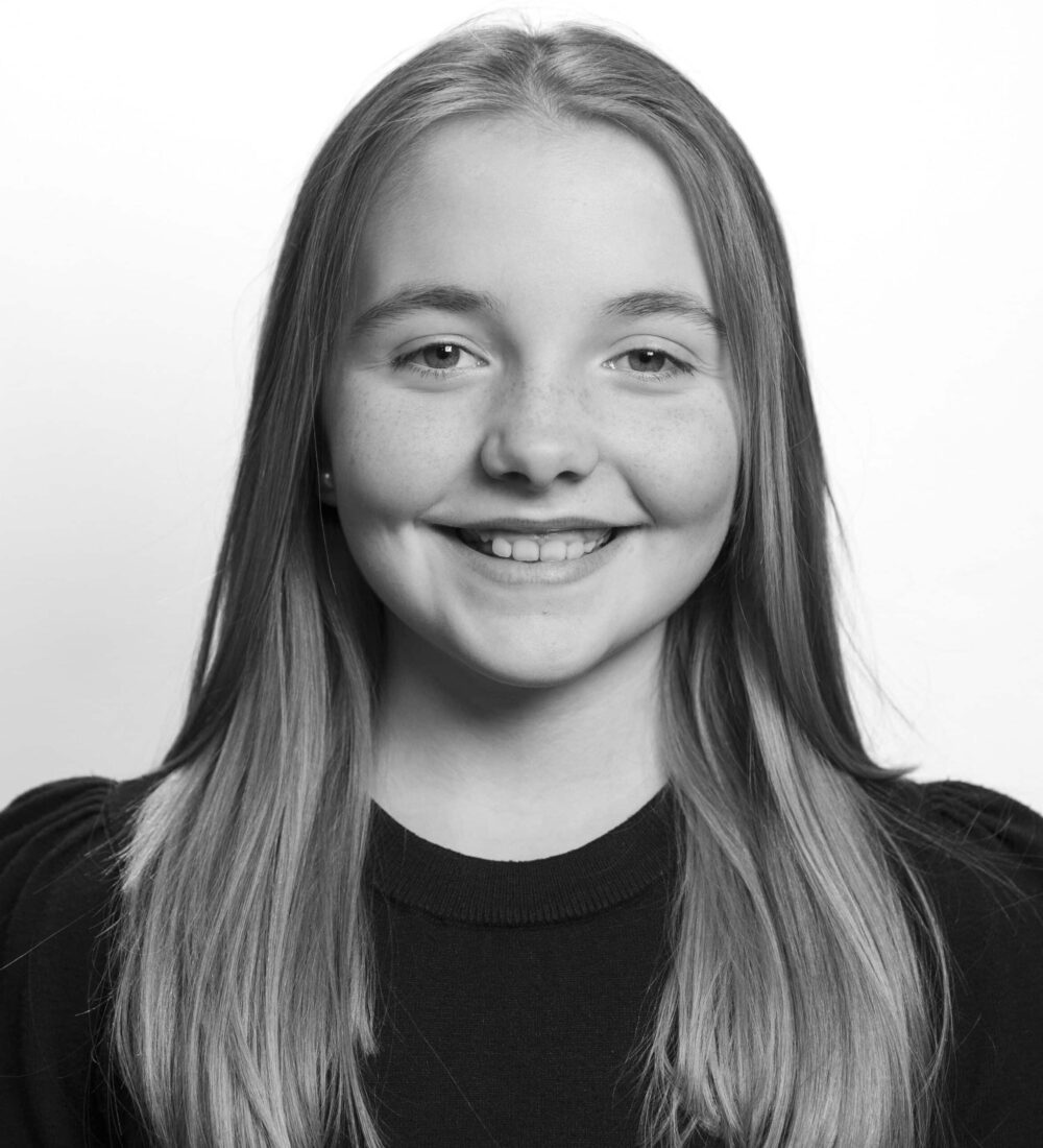 Svart-hvitt portrett av en ung jente med langt, glatt hår som smiler til kameraet mot en ensfarget bakgrunn.