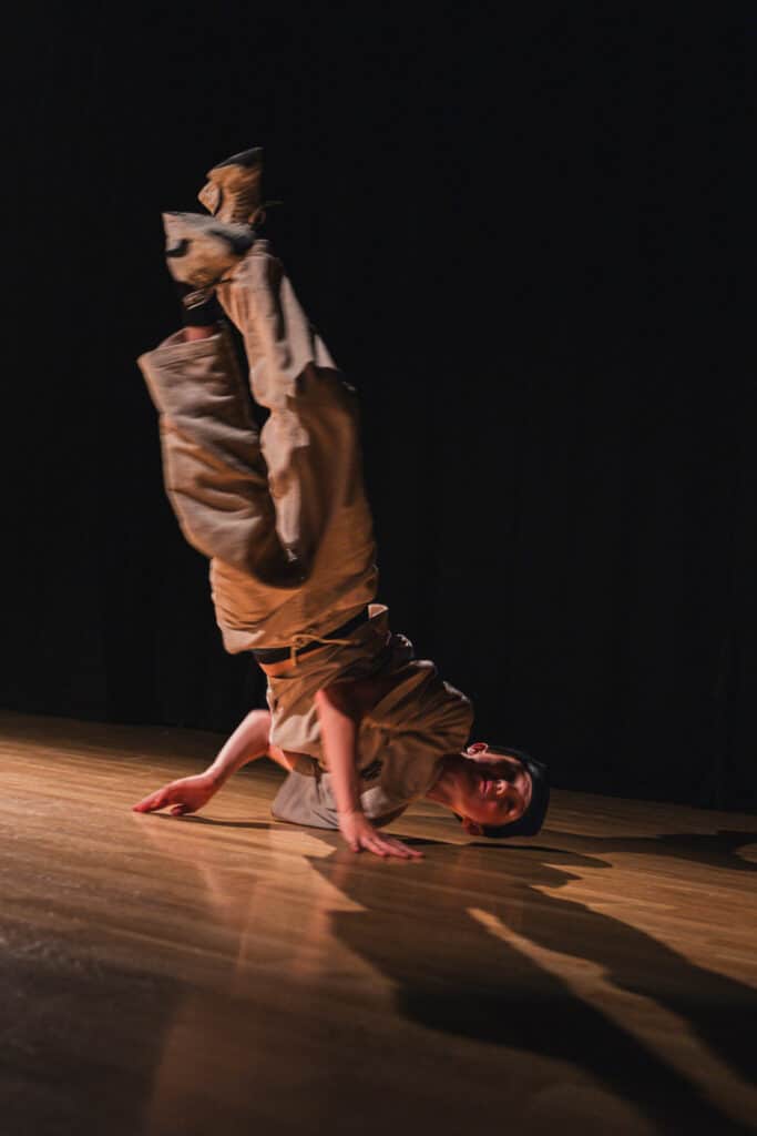 En danser i løse klær utfører en breakdance-bevegelse på scenen, balanserende på én hånd med bena løftet og bøyd, mot en mørk bakgrunn.
