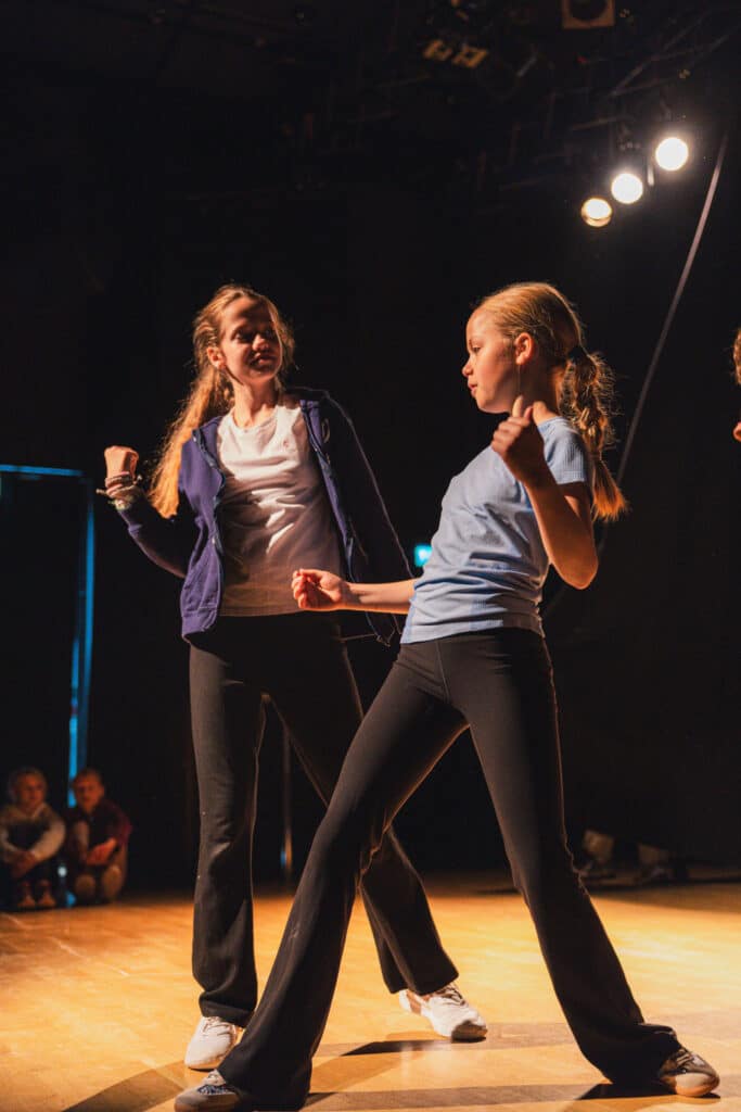 To jenter i fritidsklær danser på scenen i sterkt lys, mens noen få sittende mennesker er synlige i den dunkle bakgrunnen.