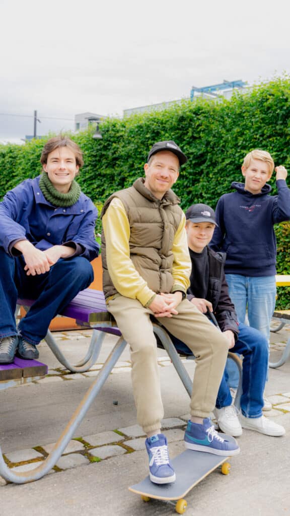 Fire personer, tre voksne og ett barn, sitter og står på og rundt et piknikbord utendørs, én person sitter på et skateboard, og i bakgrunnen skimtes grønne planter.