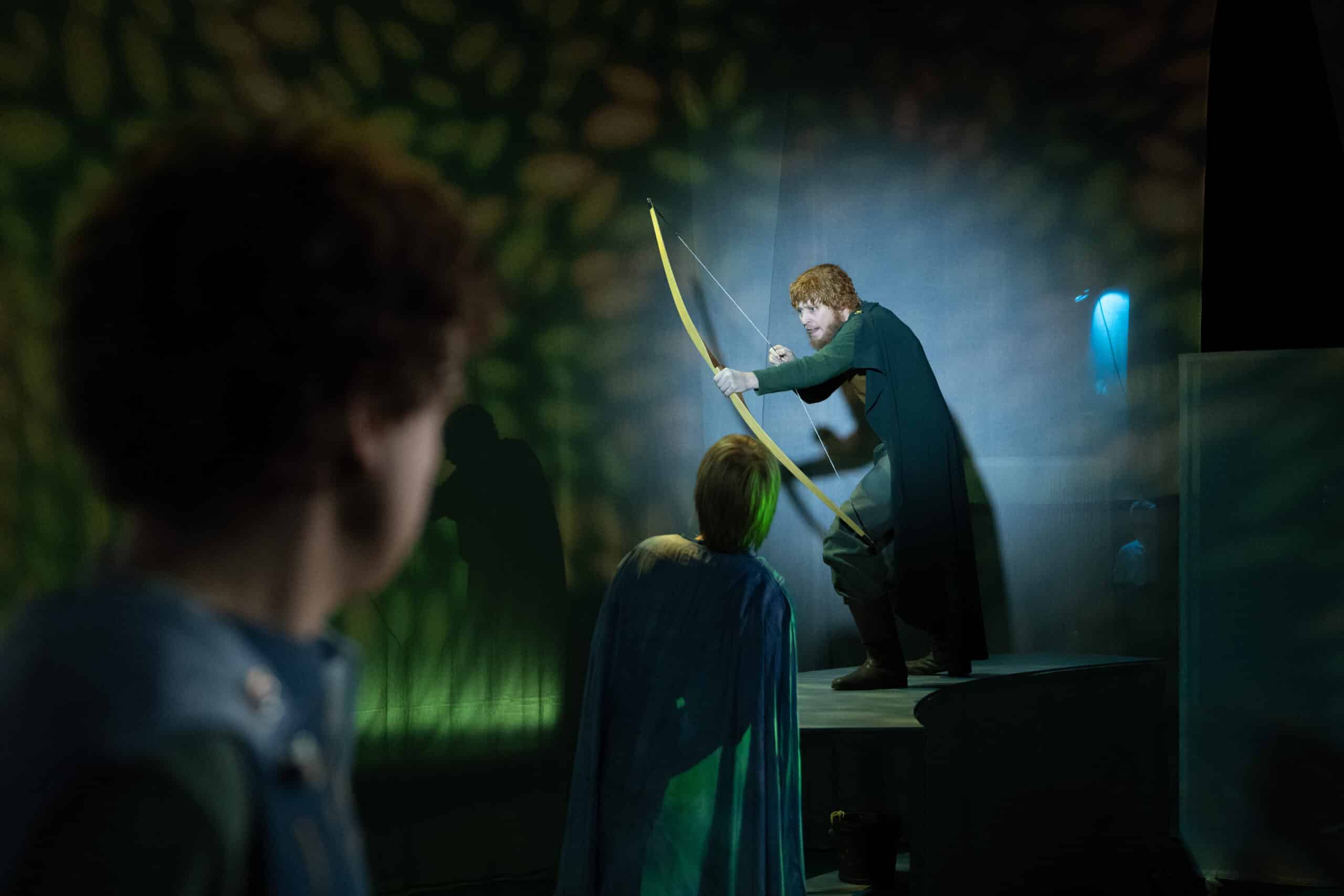 En person med krøllete hår sikter med pil og bue på en opplyst scene, mens to andre i kapper ser på i forgrunnen. Grønt og gult lys skaper en dramatisk scene.