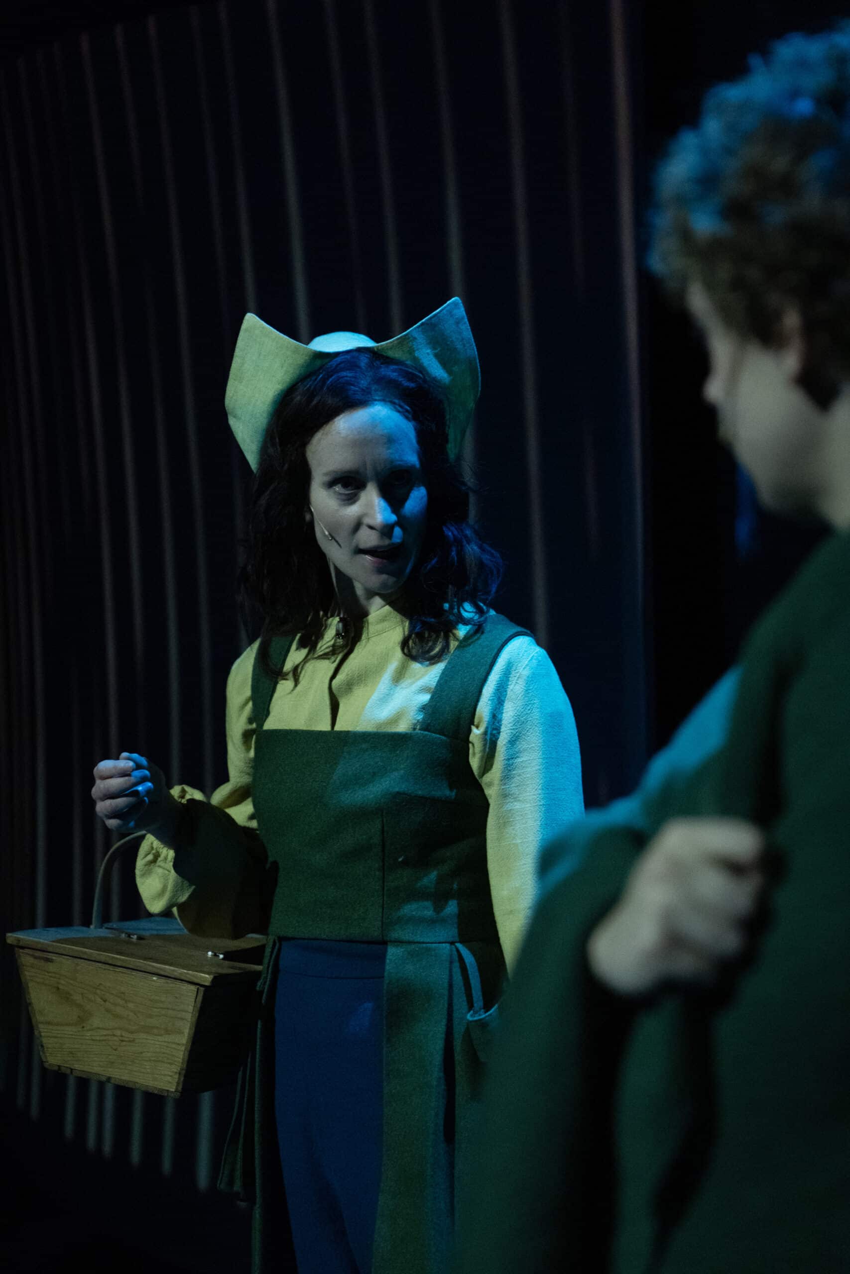 En kvinne i kostyme med grønt forkle og hodeplagg holder en kurv og snakker med en annen person på en dunkelt opplyst scene.