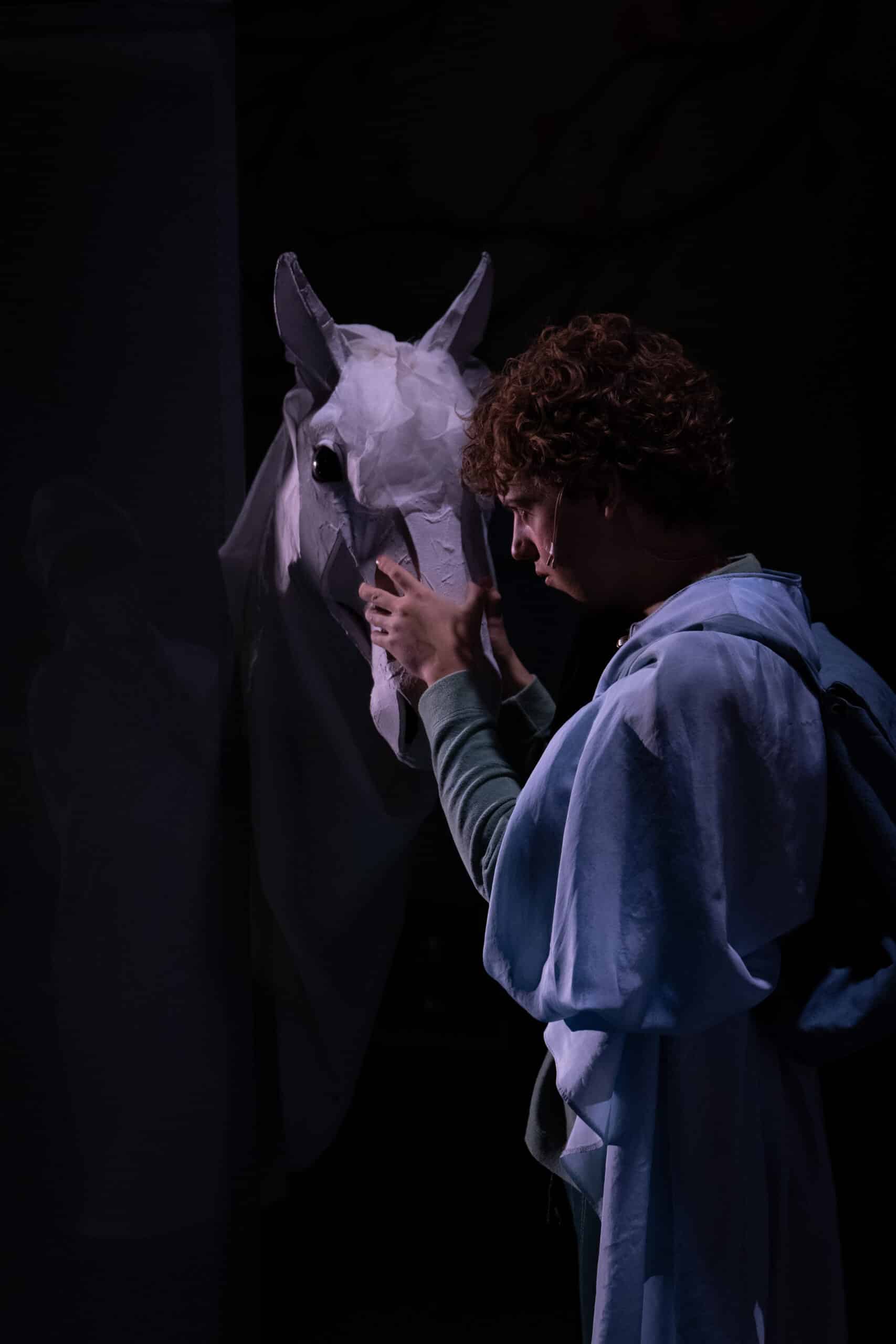 En person i blått kostyme berører forsiktig ansiktet til en hvit hestefigur på en dunkelt opplyst scene.