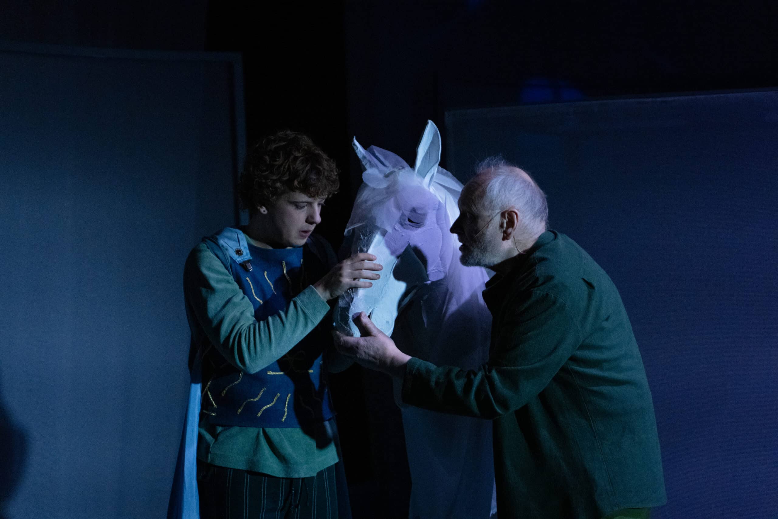 To personer interagerer med en person i et hvitt hestekostyme på en svakt opplyst scene, mens den ene forsiktig berører hestens ansikt.