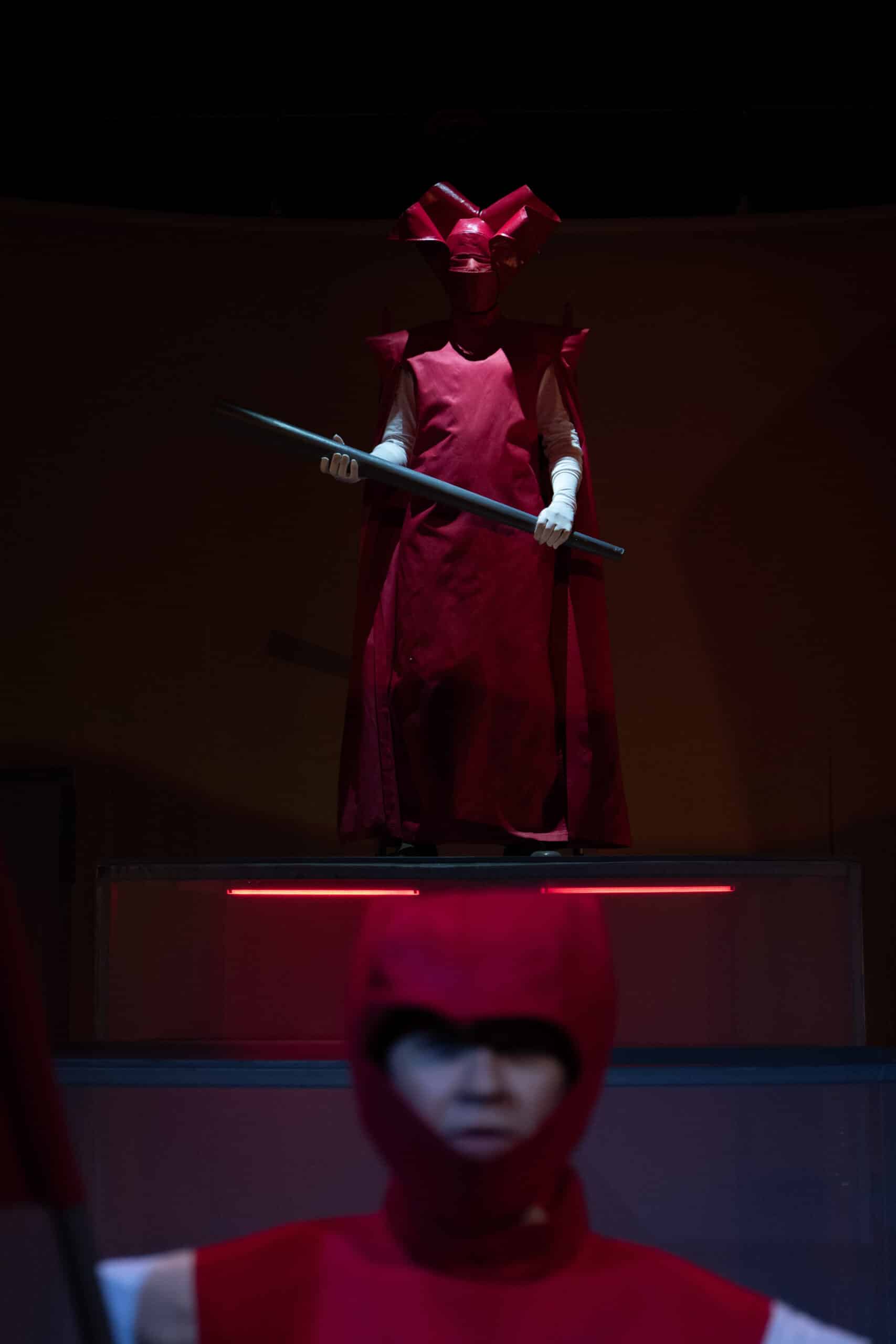 En person i dramatisk rød drakt og hodeplagg står på en forhøyet plattform og holder en stav, mens en annen person i lignende antrekk står i forgrunnen.