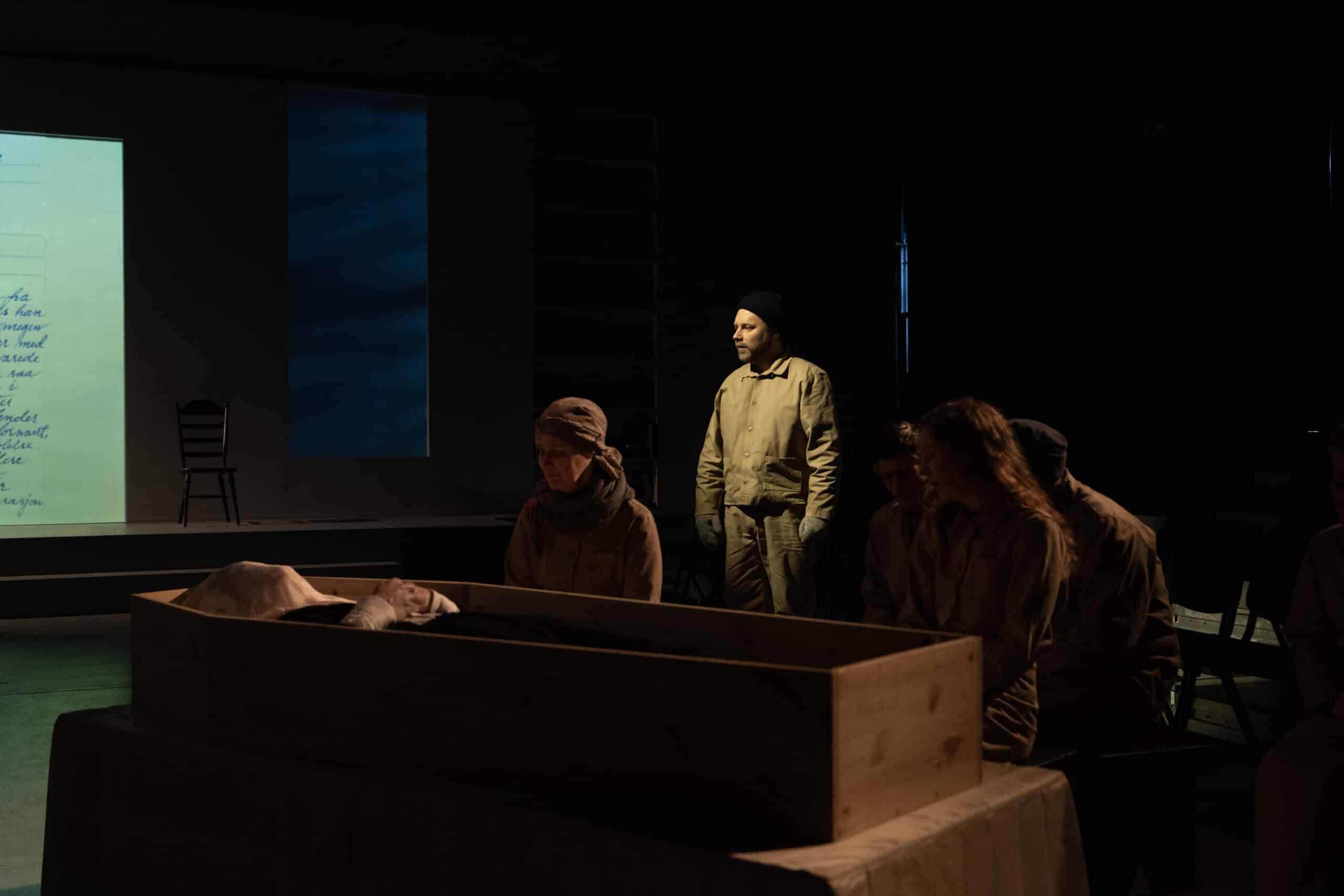 En gruppe mennesker i dunkel belysning sitter og står rundt en trekiste på scenen, med et projisert bilde og en tom stol i bakgrunnen.