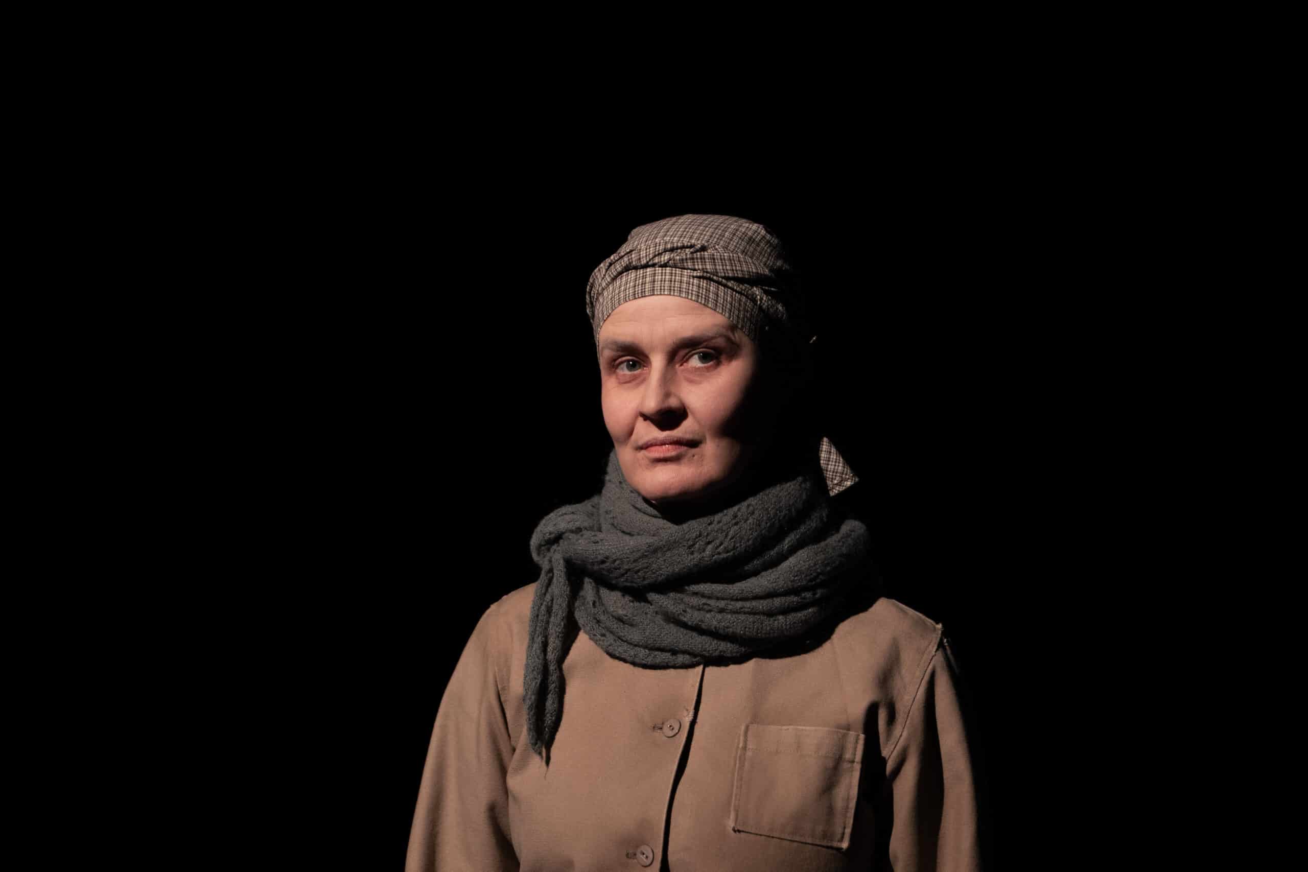 En kvinne med skaut og skjerf står mot en mørk bakgrunn, iført en nøytralfarget skjorte og ser direkte inn i kameraet.