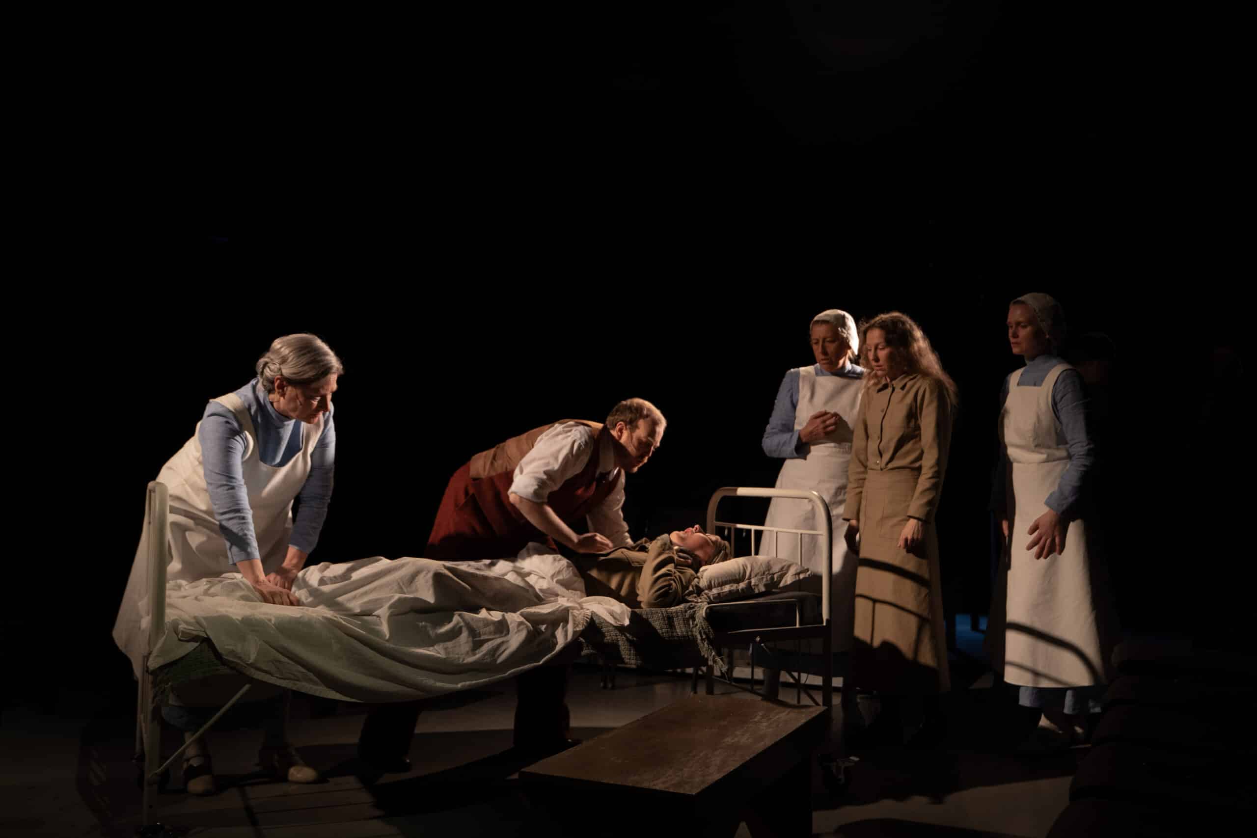 Seks personer er samlet rundt en sykehusseng; én person ligger i sengen, mens de andre står eller lener seg inn, noen av dem iført legeuniformer, i et svakt opplyst miljø.