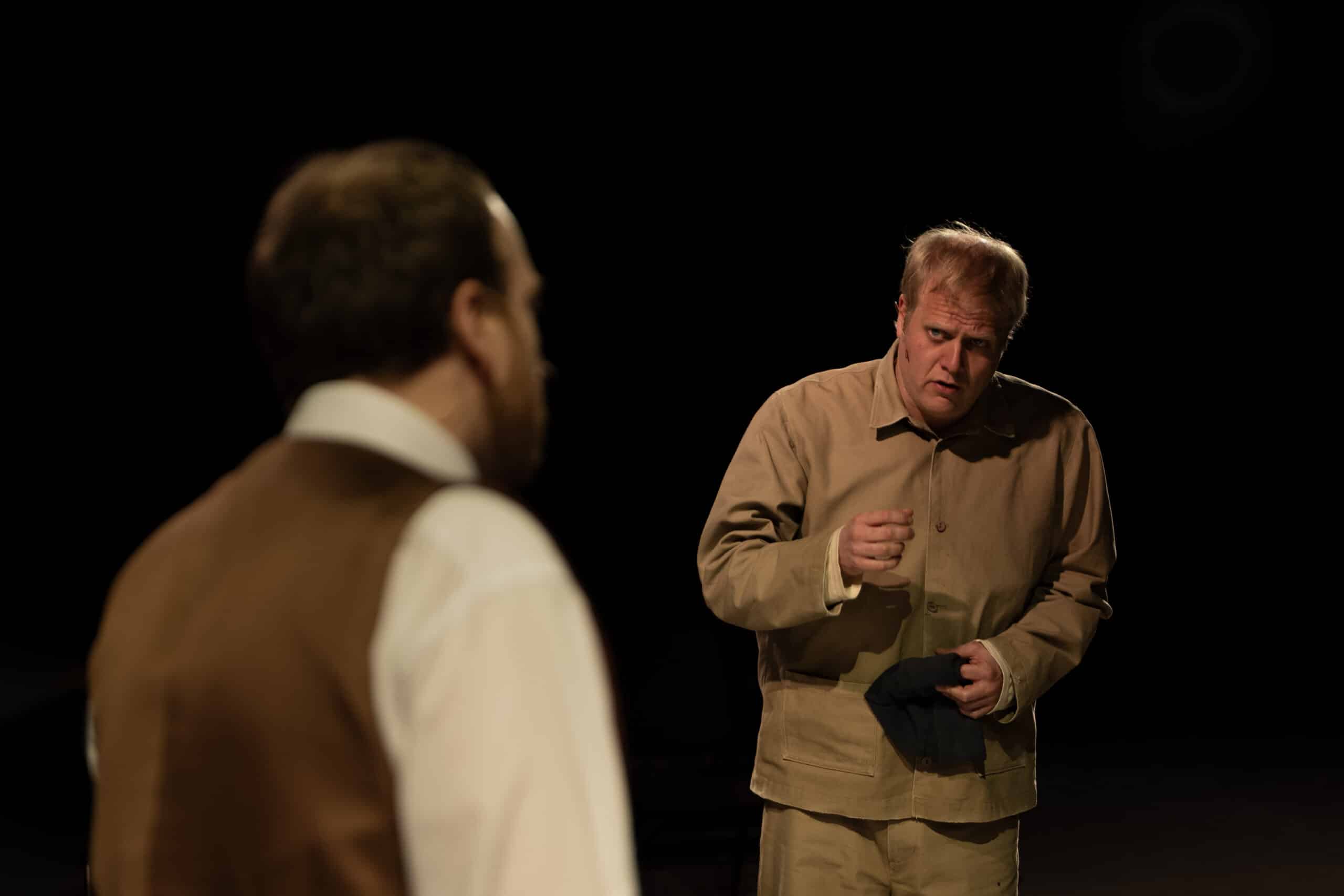 To menn står på en svakt opplyst scene; den ene i forgrunnen med ryggen til kameraet, den andre vendt fremover og gestikulerende med et alvorlig uttrykk.