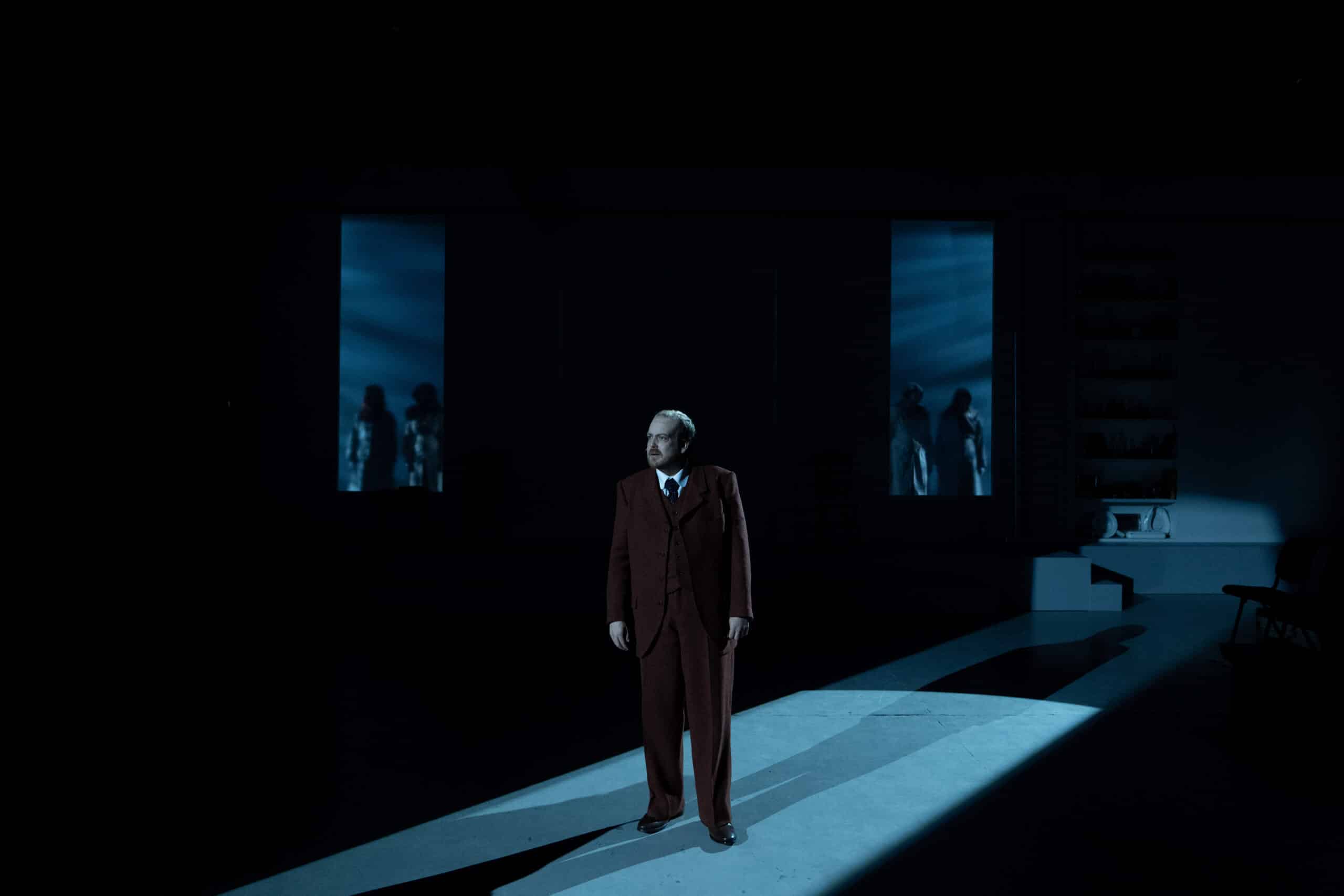 En mann i dress står alene i en spotlight på en svakt opplyst scene, med skyggerike skikkelser synlige bak to blåbelyste vinduer i bakgrunnen.
