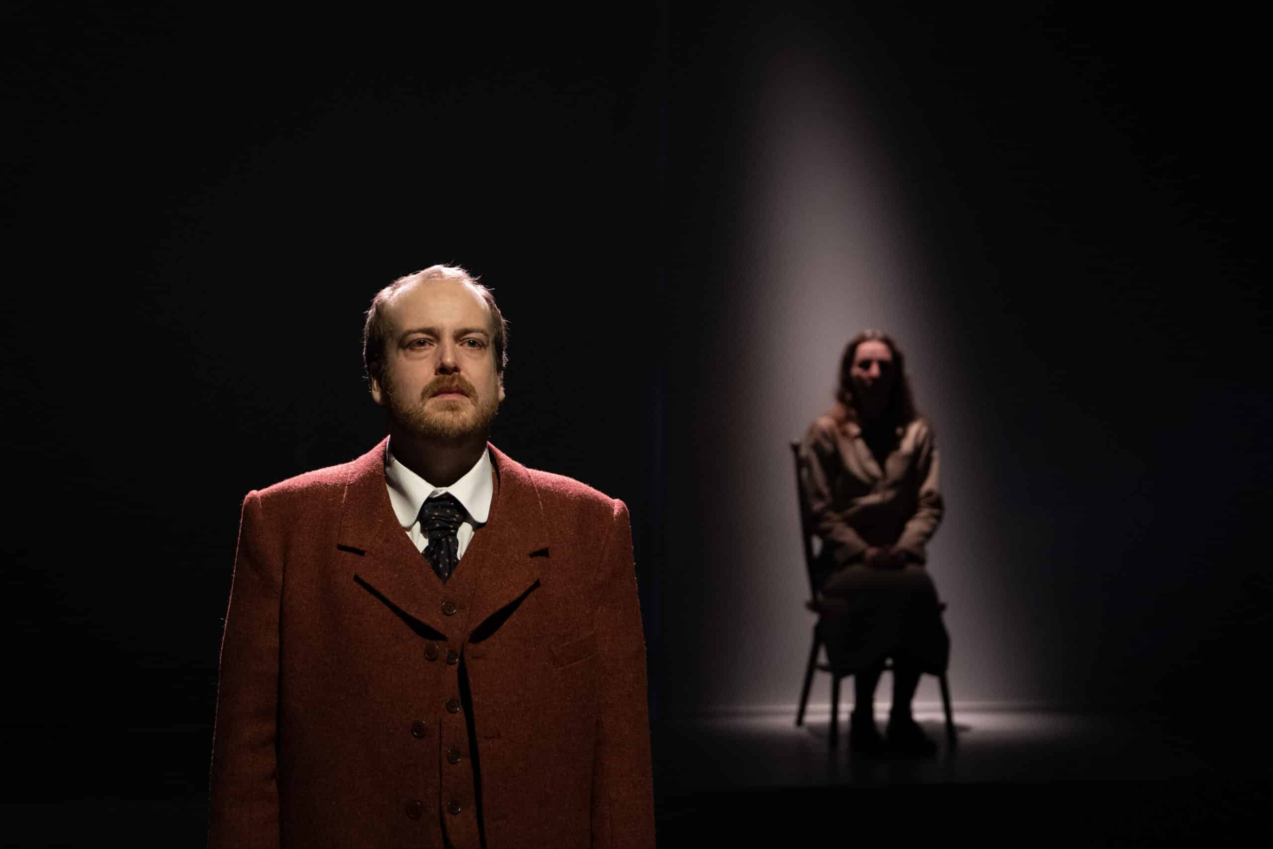 En mann i brun frakk står i forgrunnen under en spotlight, mens en kvinne sitter på en stol i bakgrunnen, også belyst av en fokusert belysning.