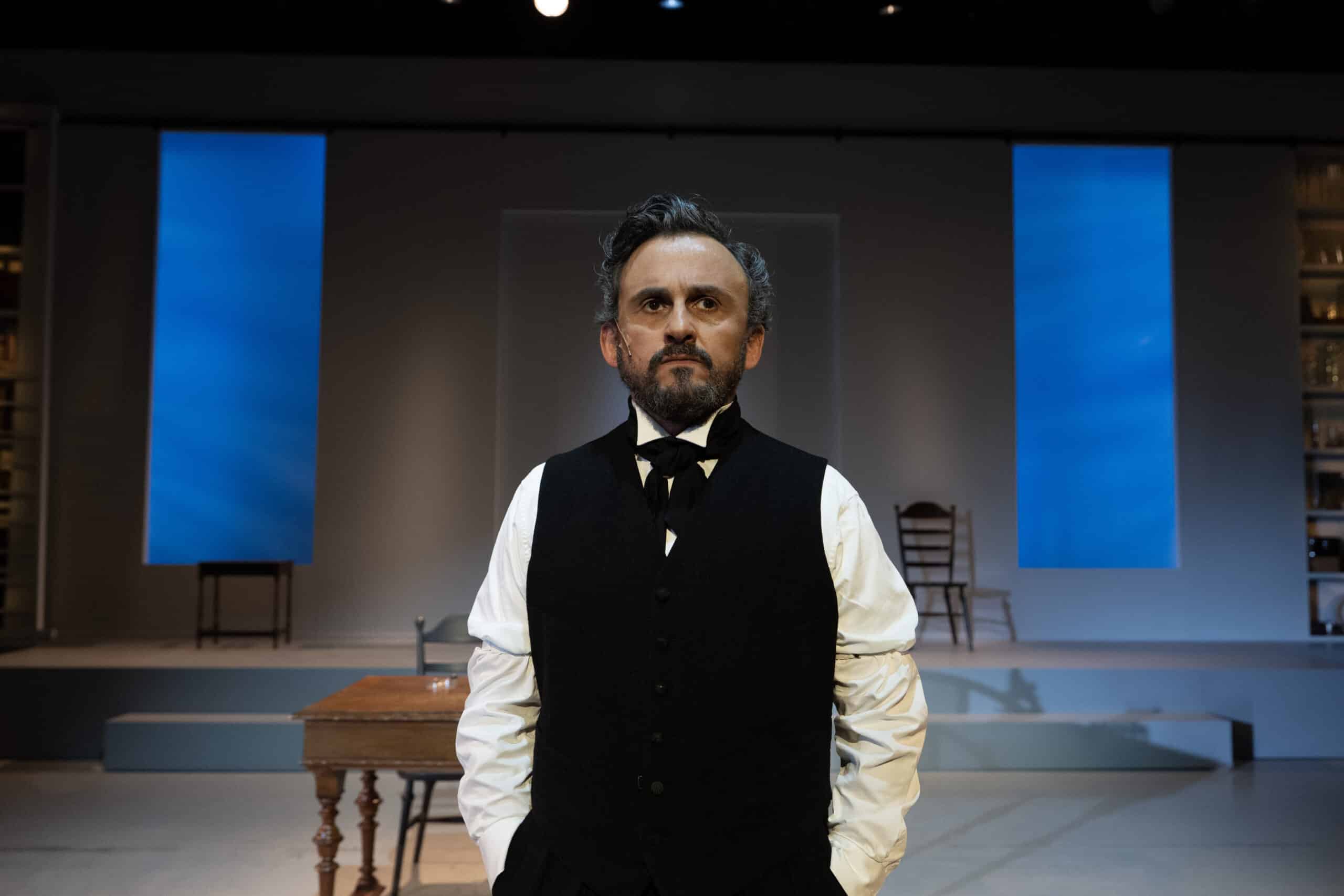 En mann i tidsriktig kostyme står på en minimalistisk scene med blå opplyste paneler, et trebord og stoler i bakgrunnen.