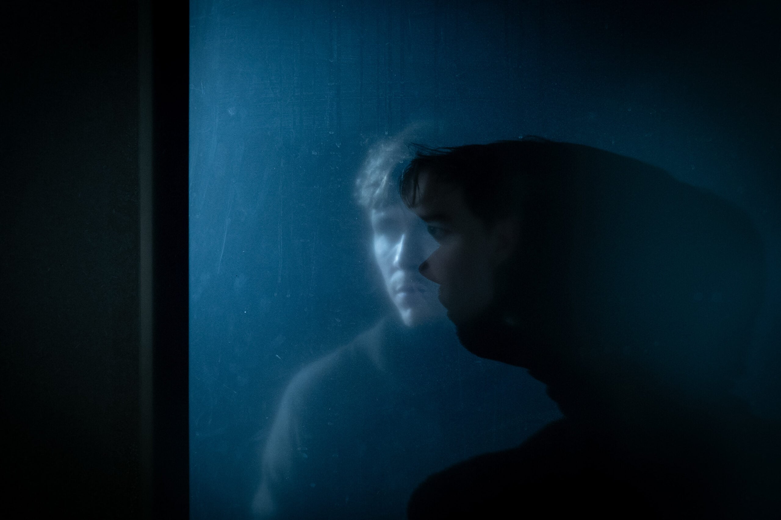 En person i et mørkt rom står vendt mot en reflekterende, blåbelyst flate, der både ansiktet og en svak refleksjon er synlig.