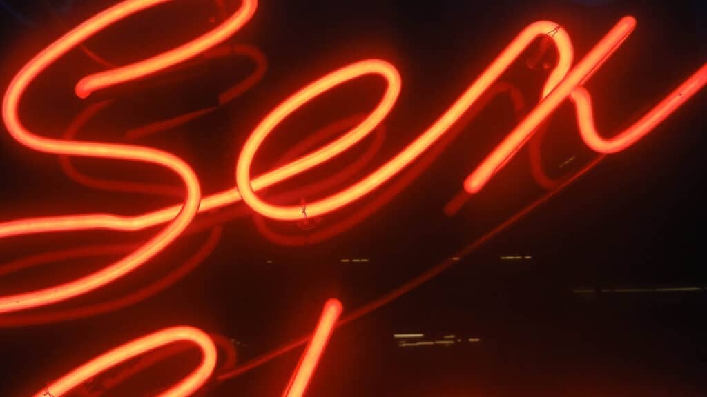 Nærbilde av et skilt med rødt neonlys som danner ordet «sex», som er delvis synlig, mot en mørk bakgrunn.