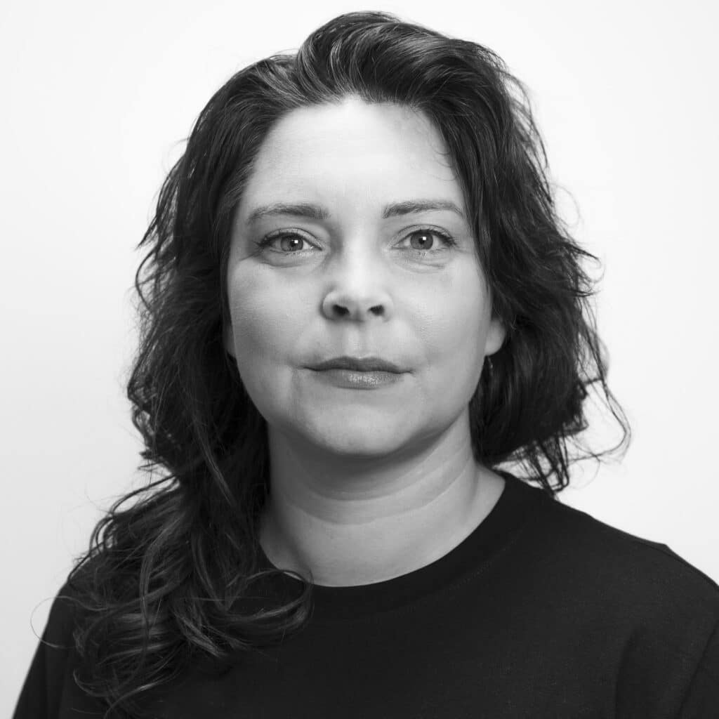 Svart-hvitt portrett av en kvinne med bølget hår og mørk skjorte, som ser direkte inn i kameraet med et nøytralt uttrykk mot en ensfarget bakgrunn.