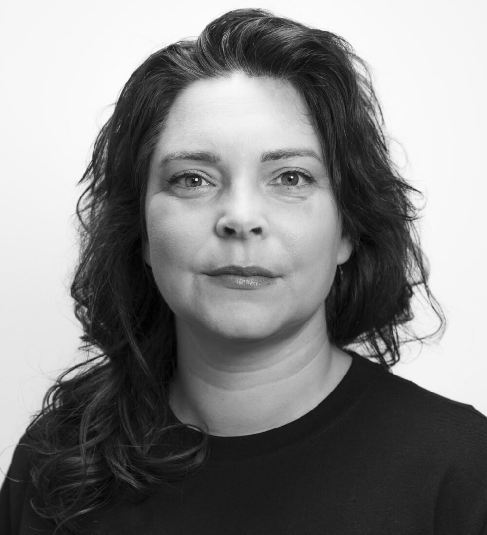 Svart-hvitt portrett av en kvinne med bølget hår og mørk skjorte, som ser direkte inn i kameraet med et nøytralt uttrykk mot en ensfarget bakgrunn.