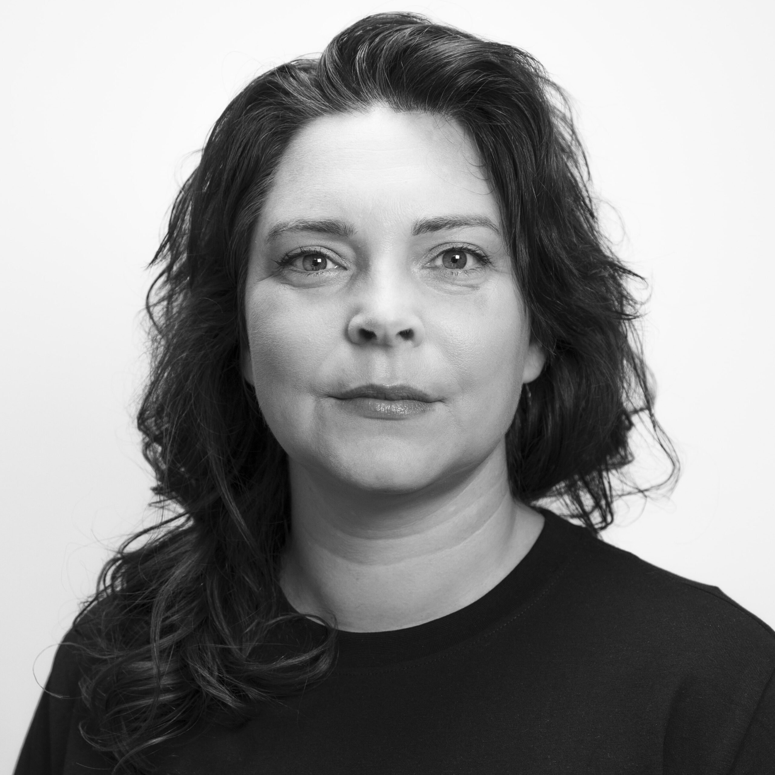 Svart-hvitt portrett av en kvinne med bølget hår og mørk skjorte, som ser direkte inn i kameraet med et nøytralt uttrykk mot en ensfarget bakgrunn.