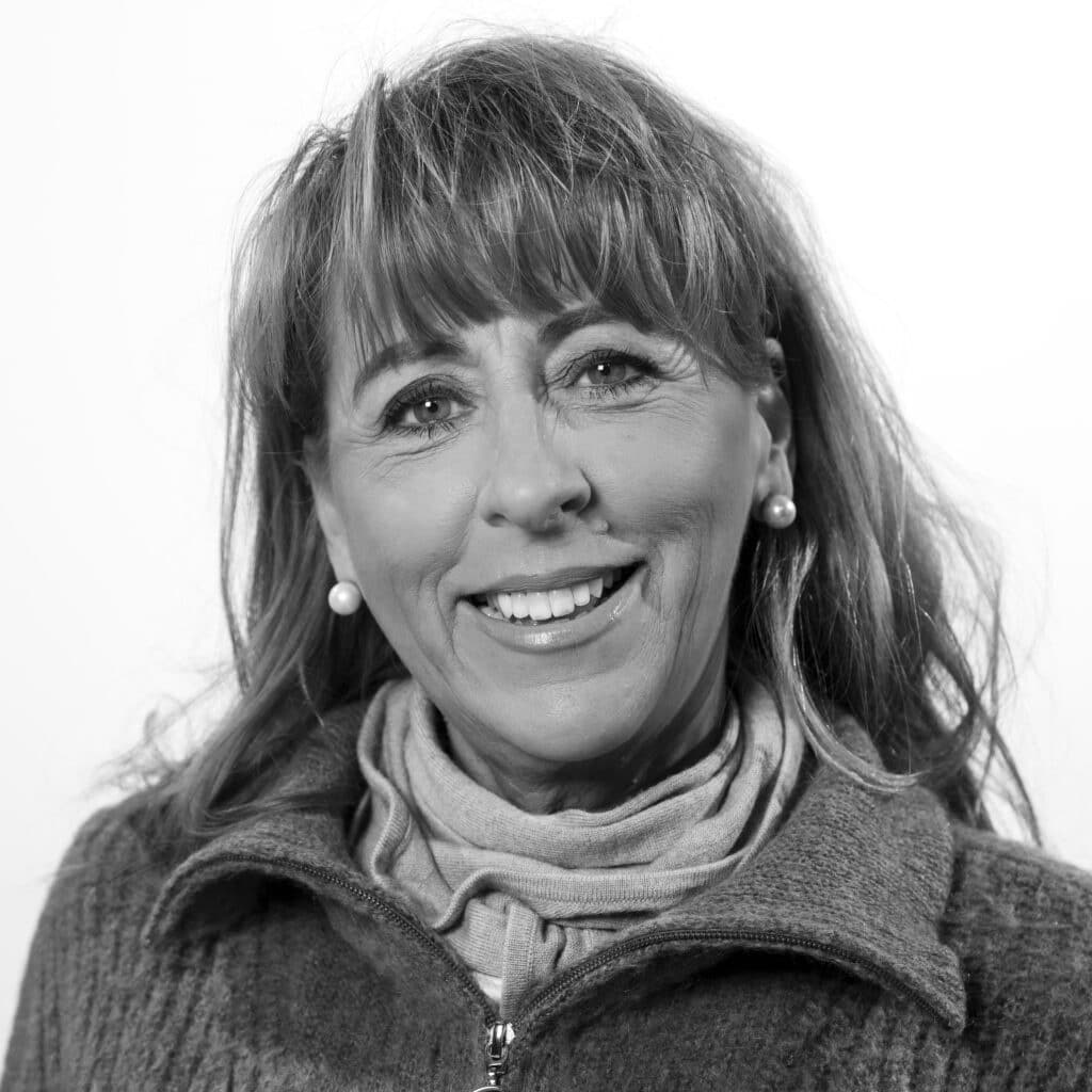 En middelaldrende kvinne med langt hår og lugg, iført genser med glidelås og skjerf, smiler til kameraet i et svart-hvitt-portrett.