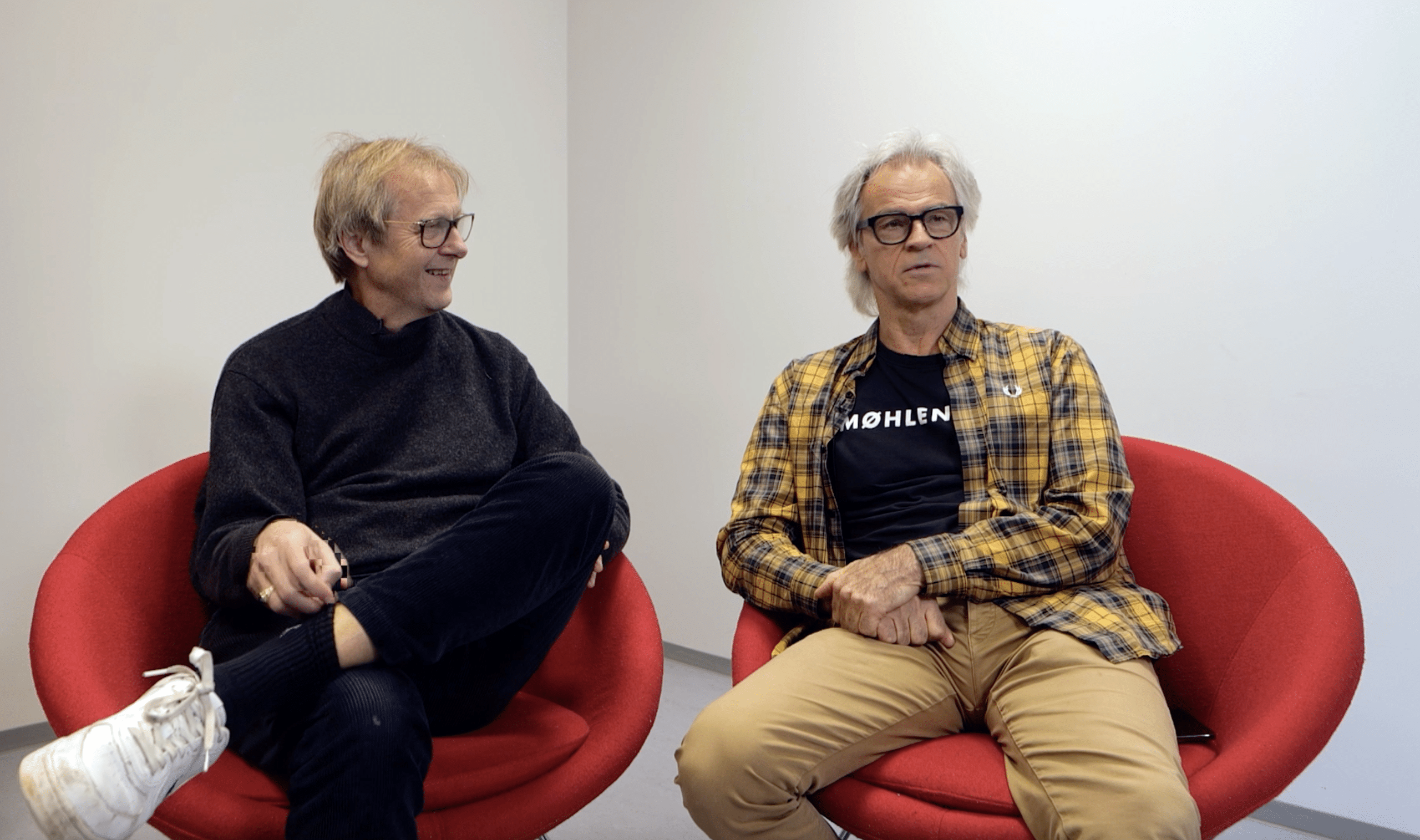 To eldre menn sitter i røde stoler mot en ensfarget vegg. Den ene har mørk genser og briller, den andre har gulrutete skjorte, T-skjorte og briller. Begge ser mot hverandre.