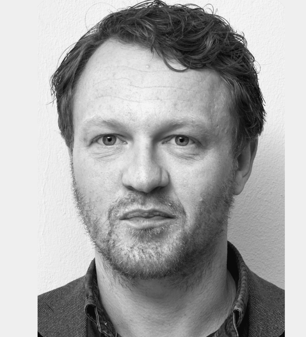 Svart-hvitt nærbilde av en mann med kort, bølget hår, lyse skjeggstubber og et nøytralt ansiktsuttrykk, stående mot en ensfarget, lys bakgrunn.