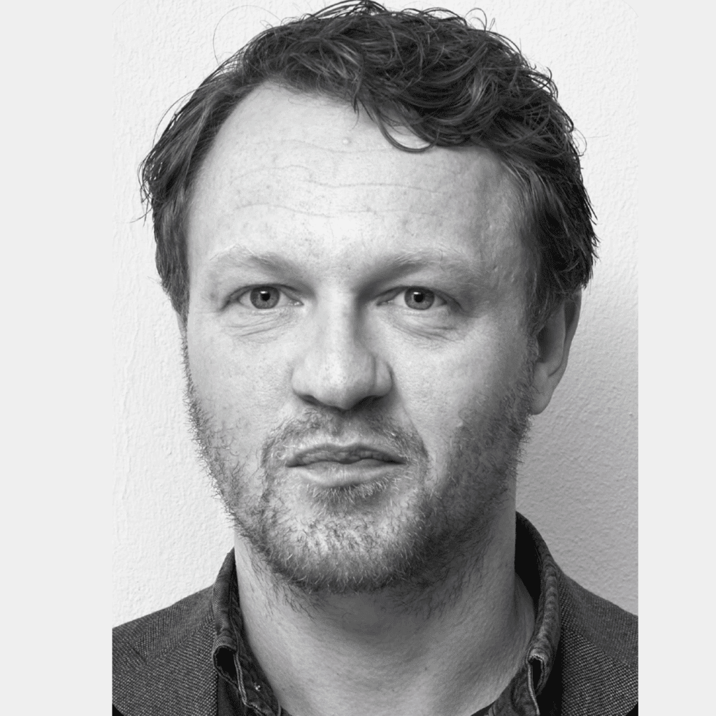 Svart-hvitt nærbilde av en mann med kort, bølget hår, lyse skjeggstubber og et nøytralt ansiktsuttrykk, stående mot en ensfarget, lys bakgrunn.