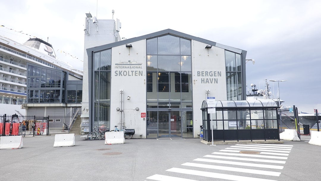 Bygning i glass og betong merket "Internasjonal Skolten Bergen Havn" ved en havn, med et skip ved kai til venstre og en fotgjengerovergang foran.