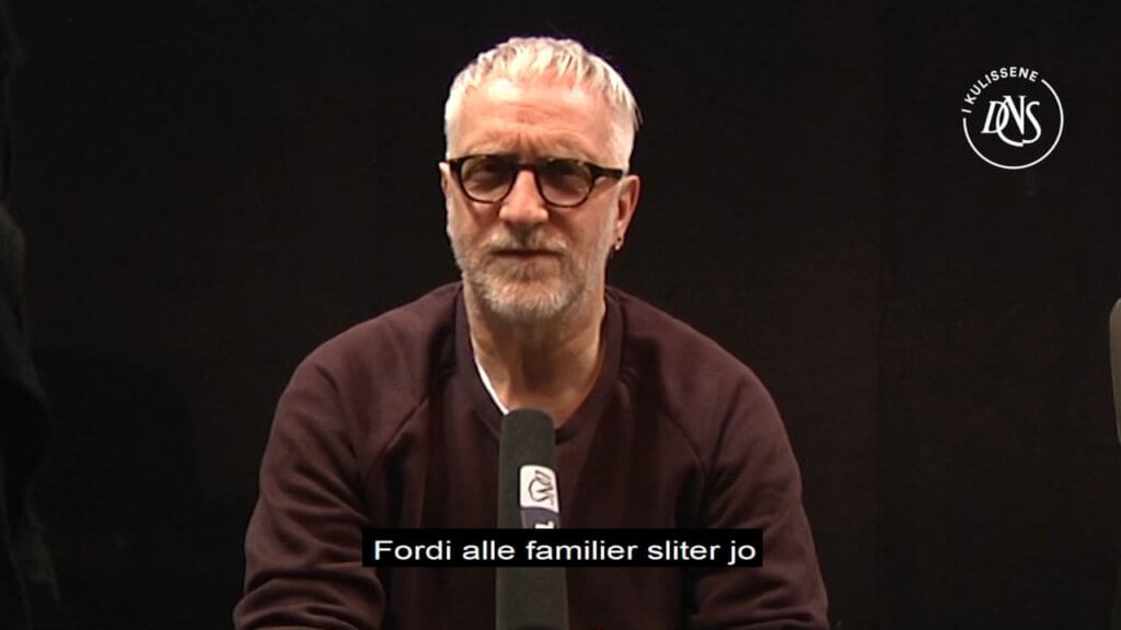 Mann med grått hår og briller sitter foran en mikrofon.