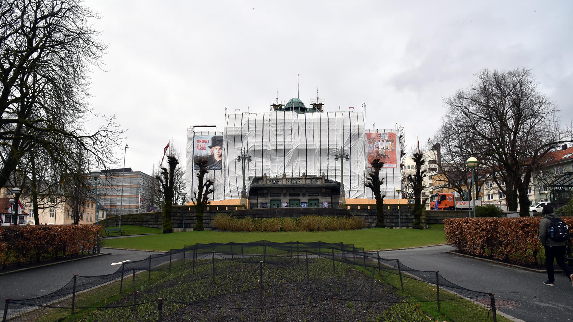 En historisk bygning som er under oppussing, dekket med stillaser og et beskyttende nett, med fotgjengere som går forbi i parklignende omgivelser.