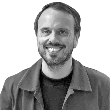 En mann med kort hår og skjegg smiler til kameraet, iført mørk skjorte og jakke, mot en enkel, hvit bakgrunn.