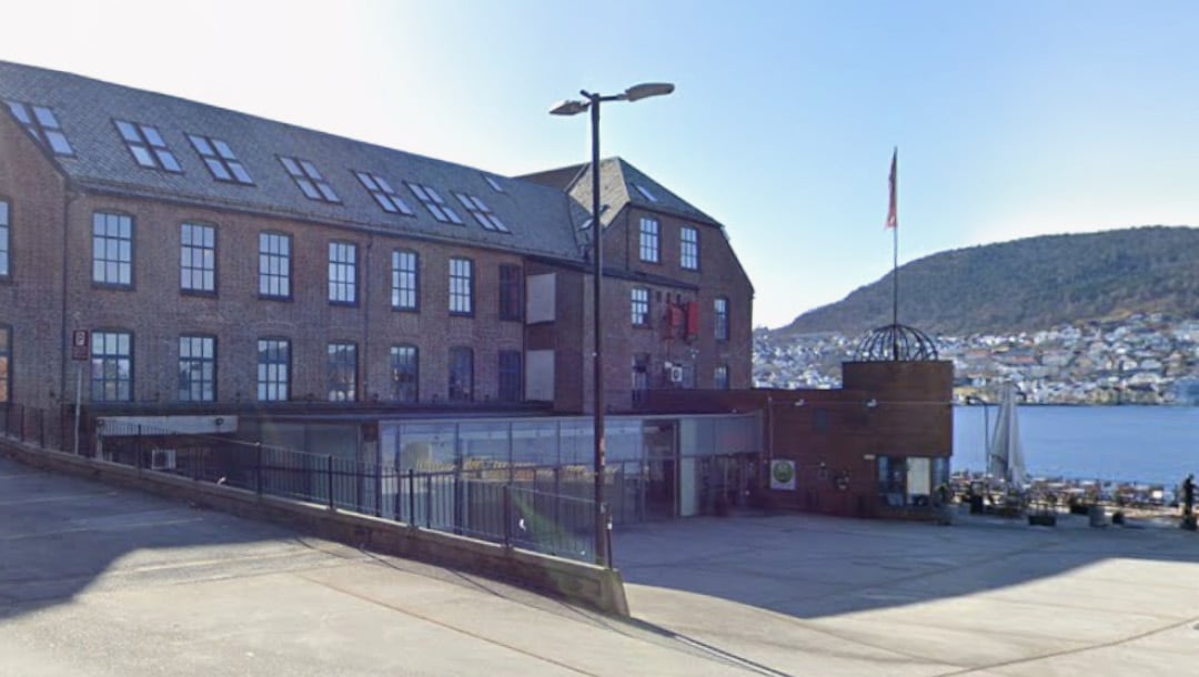 En mursteinsbygning med store vinduer og uteservering har utsikt over vannet, med åser og bygninger synlige i bakgrunnen under en klar himmel.