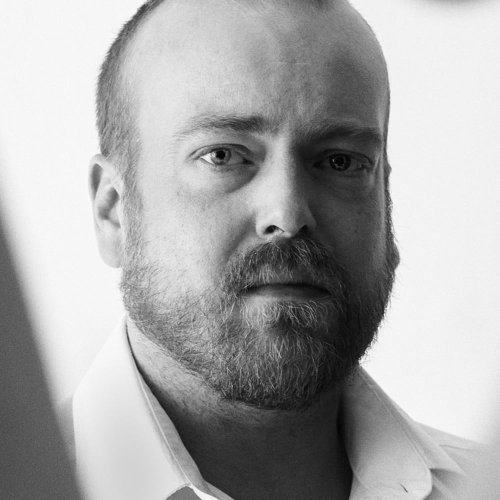 En mann med skjegg og kort hår, iført lys skjorte med krage, ser direkte inn i kameraet med et nøytralt uttrykk. Bildet er i svart-hvitt.
