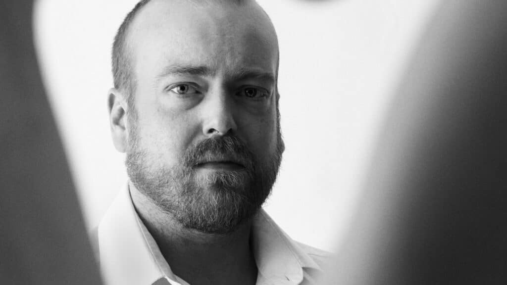 En skjeggete mann med alvorlig ansiktsuttrykk ser direkte inn i kameraet, iført hvit skjorte med krage, mot en vanlig lys bakgrunn.