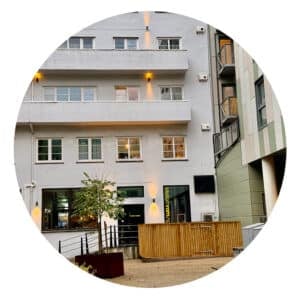 En moderne, hvit bygård med balkonger, store vinduer, et lite tre foran og et inngjerdet område ved siden av en forretningsinngang i første etasje.