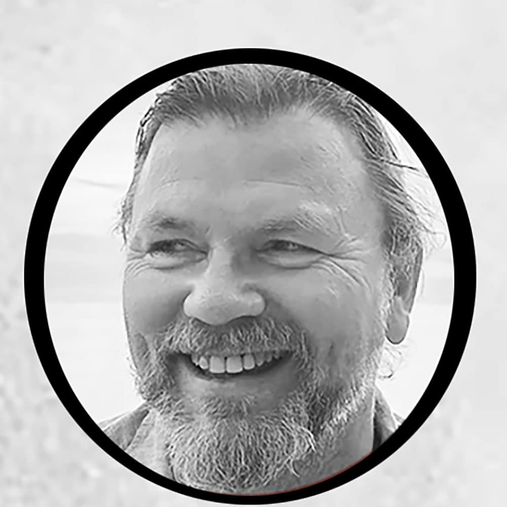 En skjeggete mann med bart smiler bredt. Bildet er i svart-hvitt og innrammet av en fet, sirkelrund ramme.