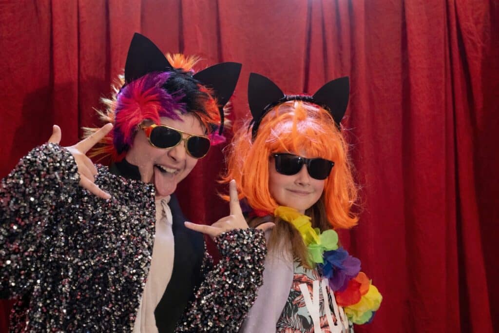 To barn foran røde gardiner iført pannebånd med katteører, fargerike parykker, solbriller og glitrende kostymer mens de poserer med lekne håndbevegelser.