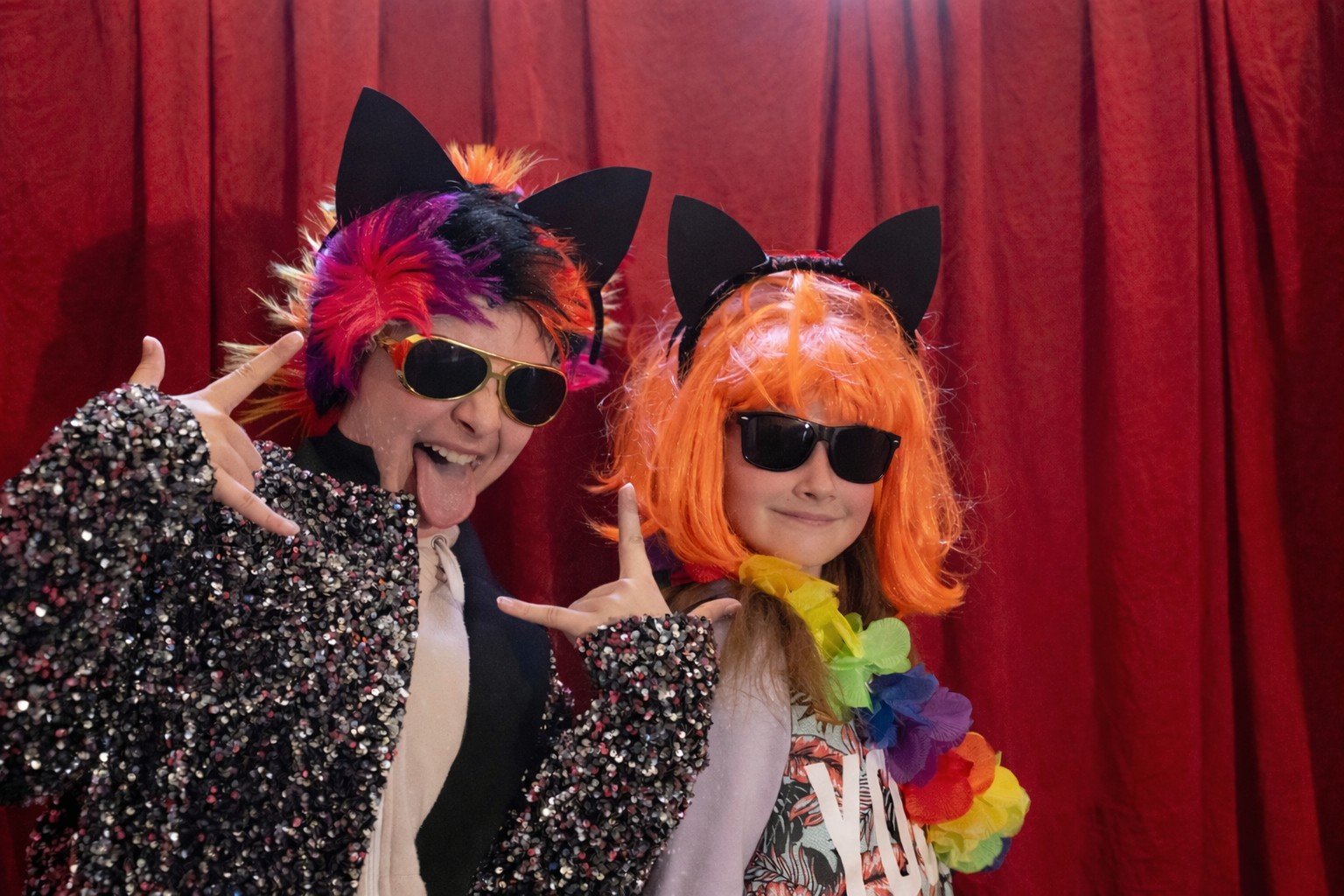 To barn foran røde gardiner iført pannebånd med katteører, fargerike parykker, solbriller og glitrende kostymer mens de poserer med lekne håndbevegelser.