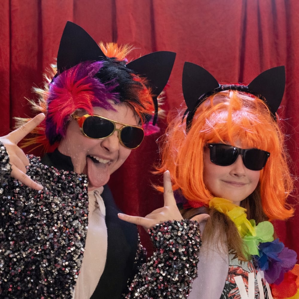 To personer i fargerike parykker, pannebånd med katteører, solbriller og paljettjakker poserer lekent foran et rødt forheng.