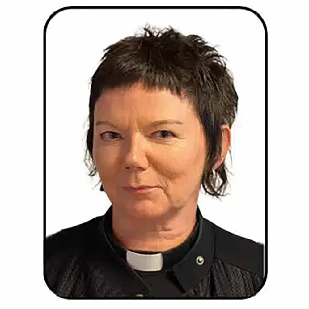 En person med kort, mørkt hår, iført svart kontorskjorte og hvit krage, ser inn i kameraet mot en hvit bakgrunn.