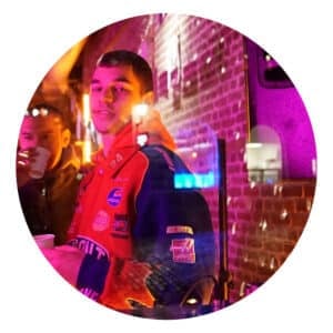 To personer står innendørs ved en murvegg, den ene iført racingjakke, opplyst av fargerike lys og refleksjoner fra et vindu i nærheten.