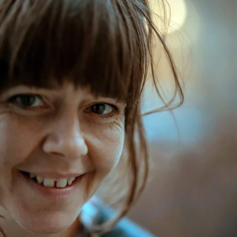 En kvinne med brunt hår og pannelugg smiler til kameraet, med en mykt uskarp bakgrunn.