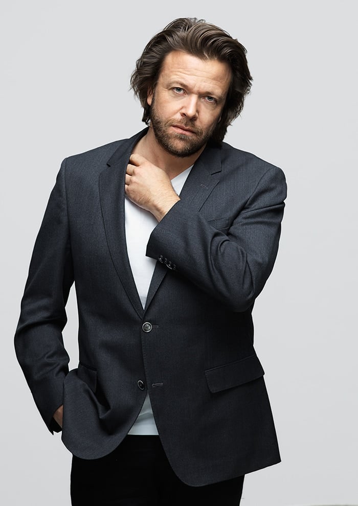 En mann med middels langt brunt hår og skjegg, kledd i en mørk blazer over en hvit skjorte, som står foran en ensfarget lysegrå bakgrunn og ser inn i kameraet med et nøytralt uttrykk.