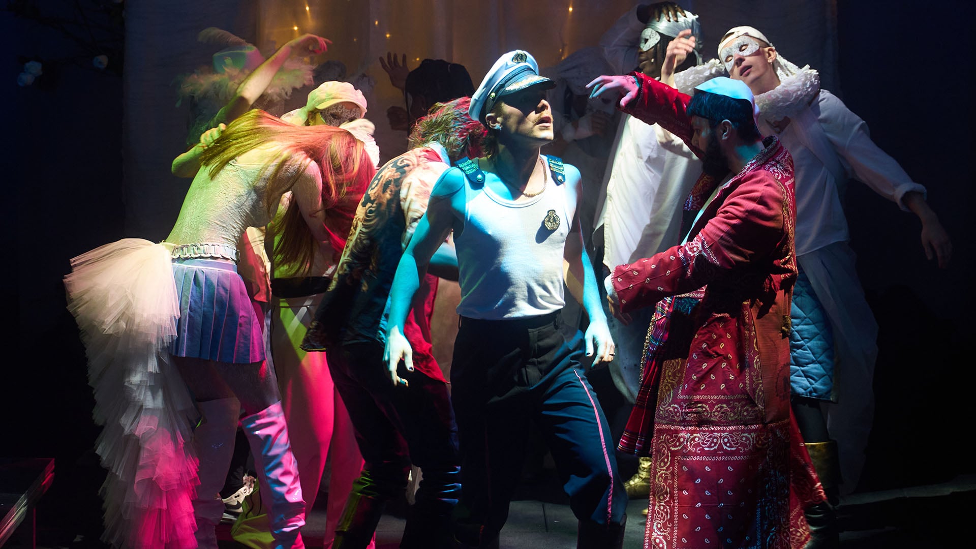 En gruppe mennesker i fargerike, teatralske kostymer danser og poserer på scenen i dramatisk belysning.