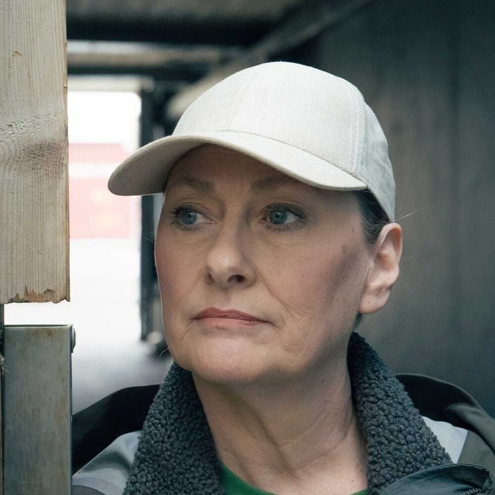 En person med en lys baseballcap og jakke står ved siden av en trepæl i en utendørs gang og ser litt til siden.