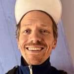 En mann iført hvit lue, marineblå jakke med glidelås og ryggsekk smiler til kameraet foran en lyselilla vegg.