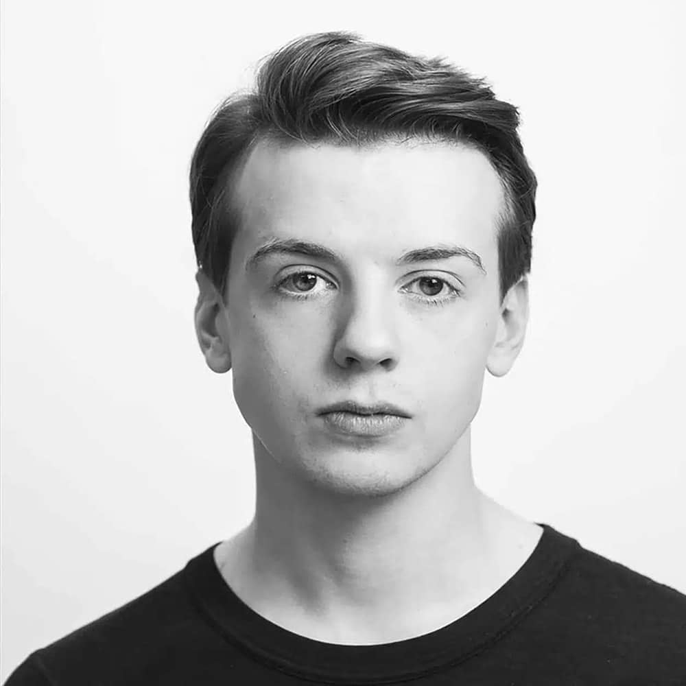 Ung person med kort, velfrisert hår og nøytralt ansiktsuttrykk, iført mørk skjorte med rund hals, fotografert mot en vanlig hvit bakgrunn.